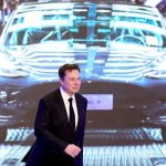 Elon Musk, gekleed in een zwart pak met een zwarte stropdas, loopt over een podium met een projectie van een auto achter hem.