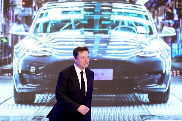 Elon Musk, gekleed in een zwart pak met een zwarte stropdas, loopt over een podium met een projectie van een auto achter hem.